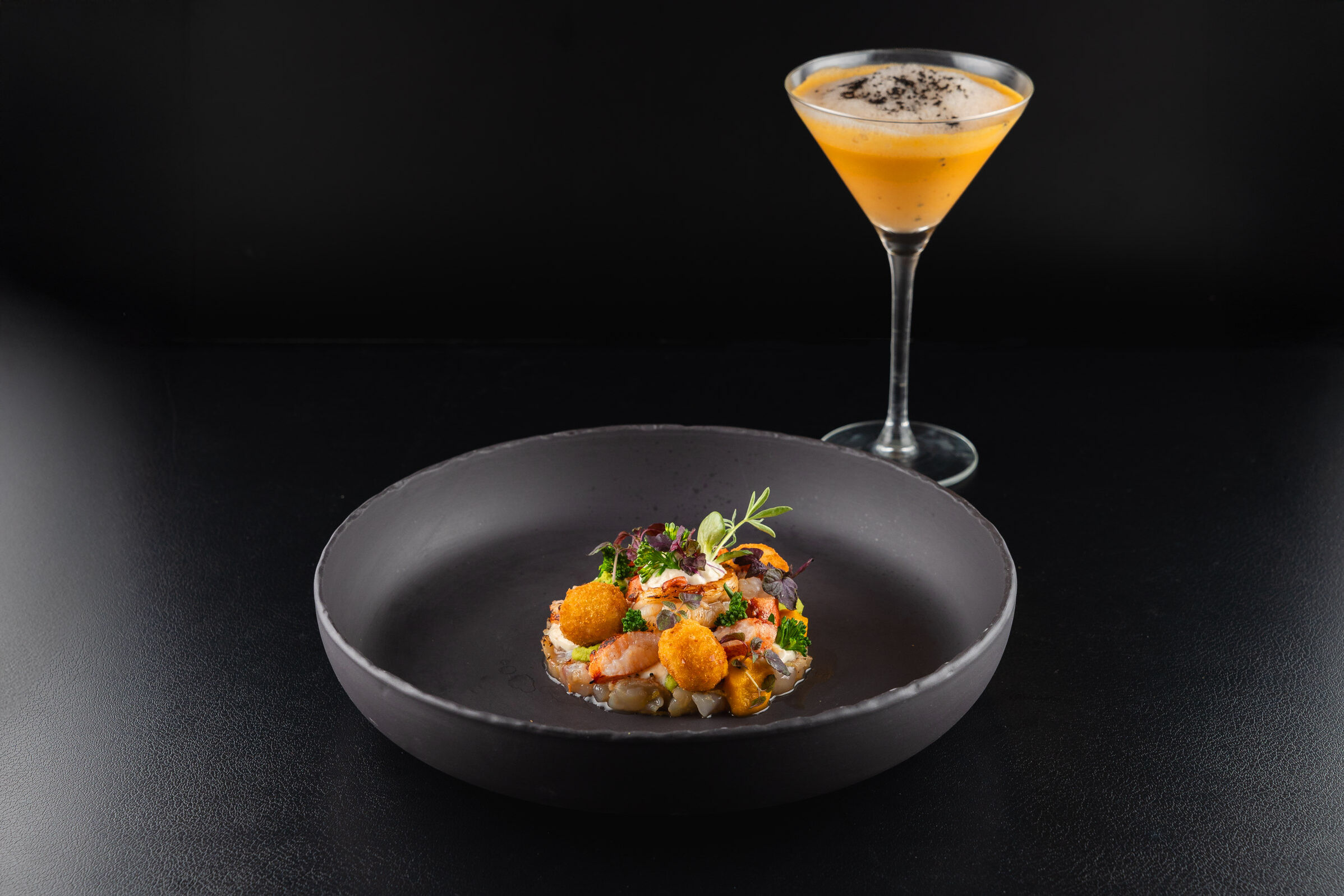 Lobster dish on a black plate, with a glass in the background.