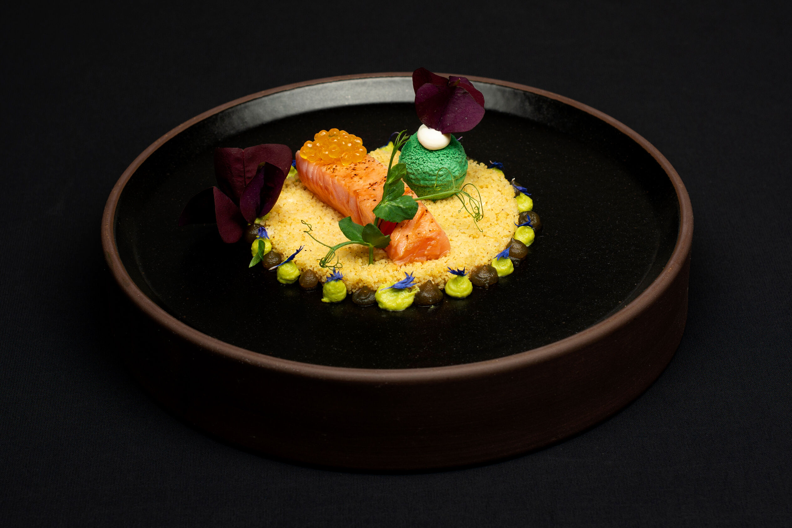Schwarzer Teller mit zarten Lachsfilets, Erbse, Minze und lockerem Couscous, elegant präsentiert.