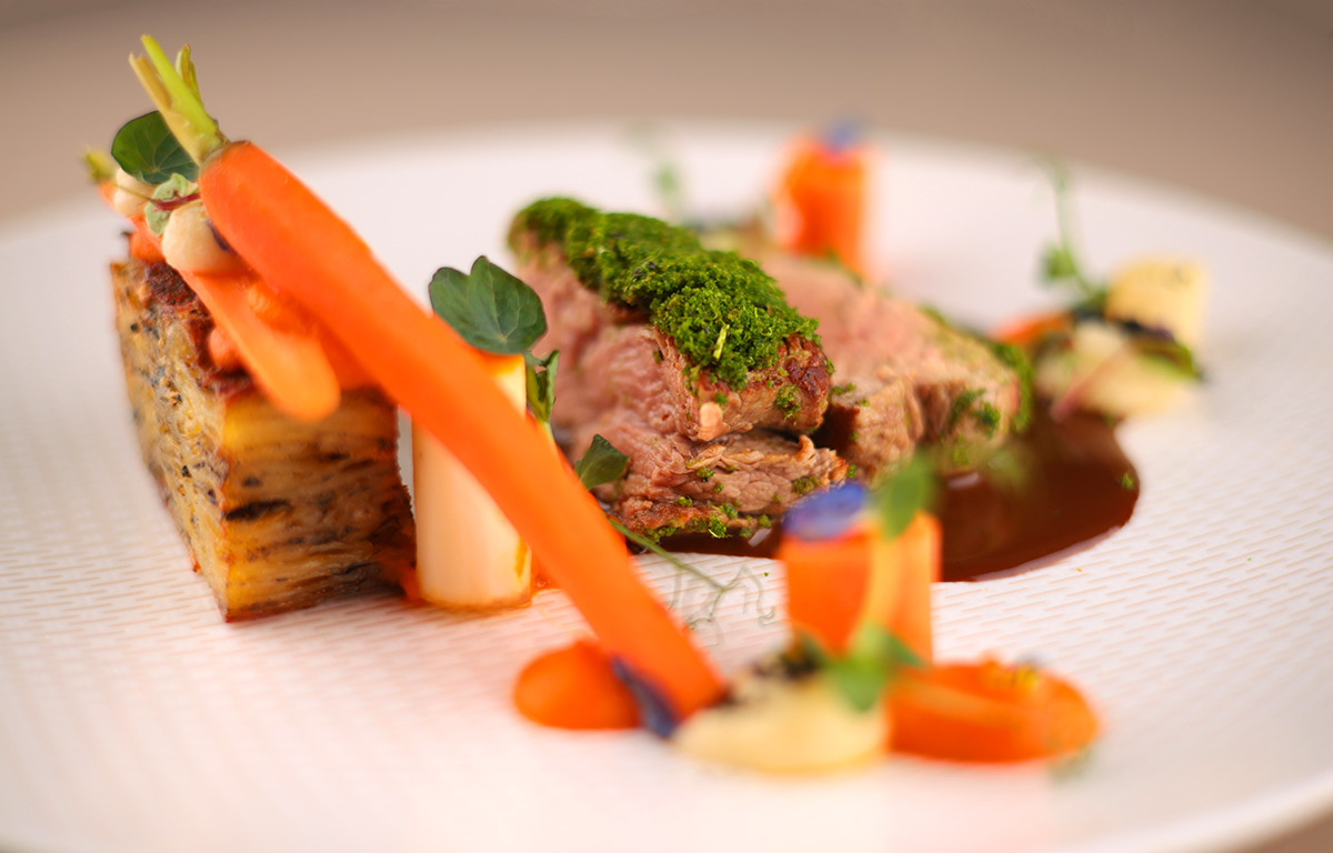 Zartes Kalbsfilet mit Kräuterkruste, orange Möhren, grüner Kruste und essbaren Blüten.