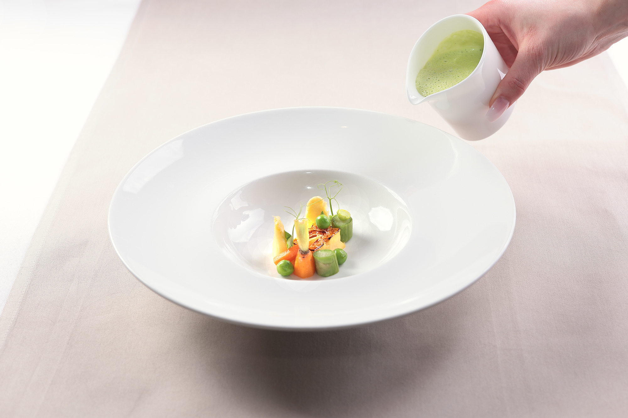 Erbsen-Velouté wird aus weißem Kännchen auf frisches Gemüse im Teller gegossen, sanft umhüllt.